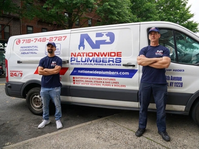 Two plumbers stand proudly in front of their company van.