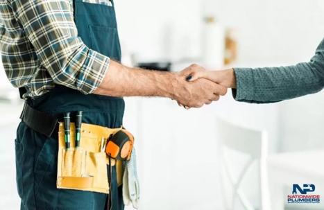 A plumber in a toolbelt shakes hands with a customer.