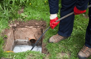 A person, wearing gloves and work boots, clears a drain with a long tool.