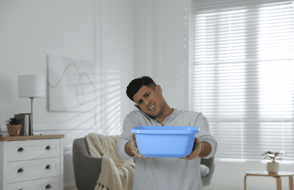 A man is holding a blue basin under a ceiling leak while on his phone.