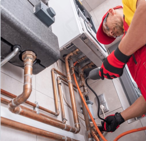 A technician in red overalls repairs a boiler with copper piping.